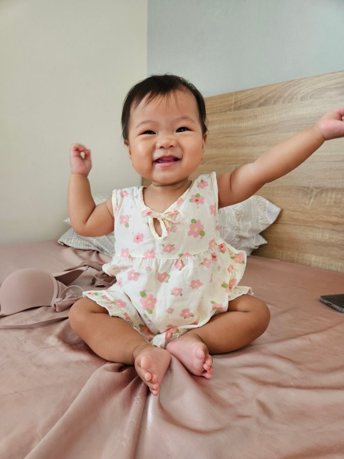 Baby in a flower outfit sitting on a bed with one arm up, smiling